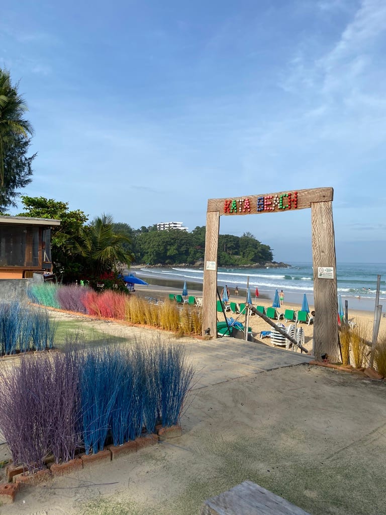 A wooden KATA BEACH archway opening onto the sand, with loungers, umbrellas and breaking surf beyond