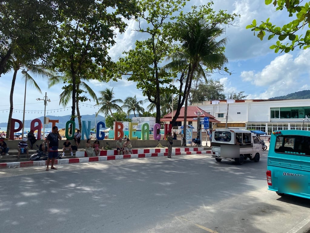 The PATONG BEACH sign on the beach road, songthaews passing in front of it and the sand just visible behind