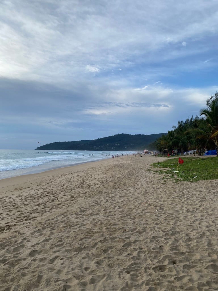 A wide beach at Karon under a grey monsoon sky, a red warning flag planted on the grass strip at the back of the sand, with breaking surf and a scattering of people standing in the water in the distance