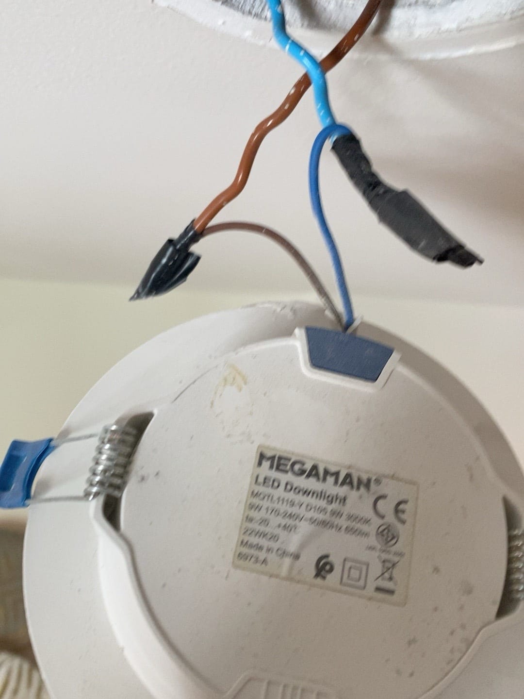 A round white LED downlight removed from a ceiling, hanging by its brown and blue wires, with the MEGAMAN label reading 9W 3000K 650lm D105 on the back