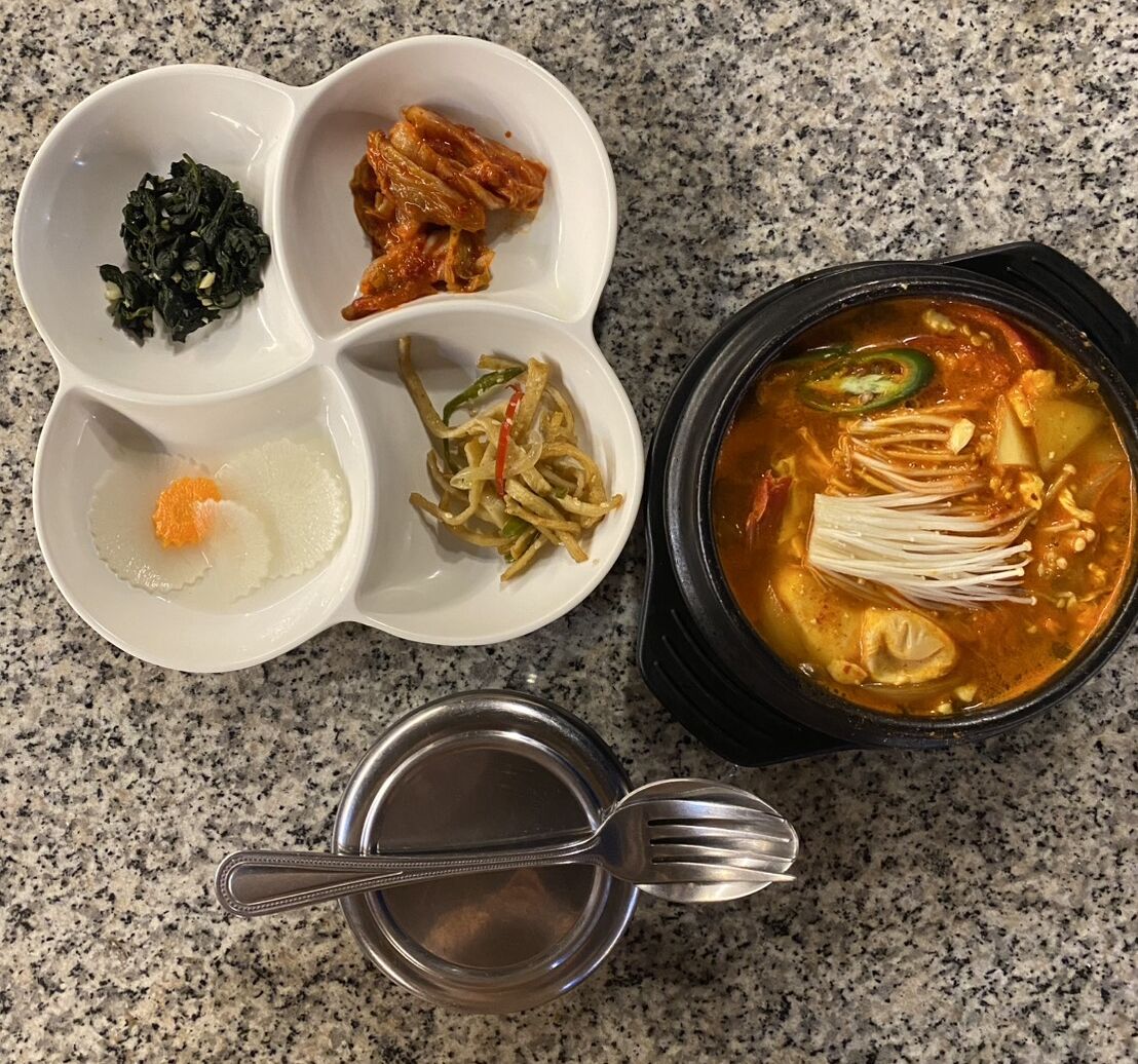 Korean sundubu jjigae in a black stone pot beside a four-compartment dish of banchan, on a granite table