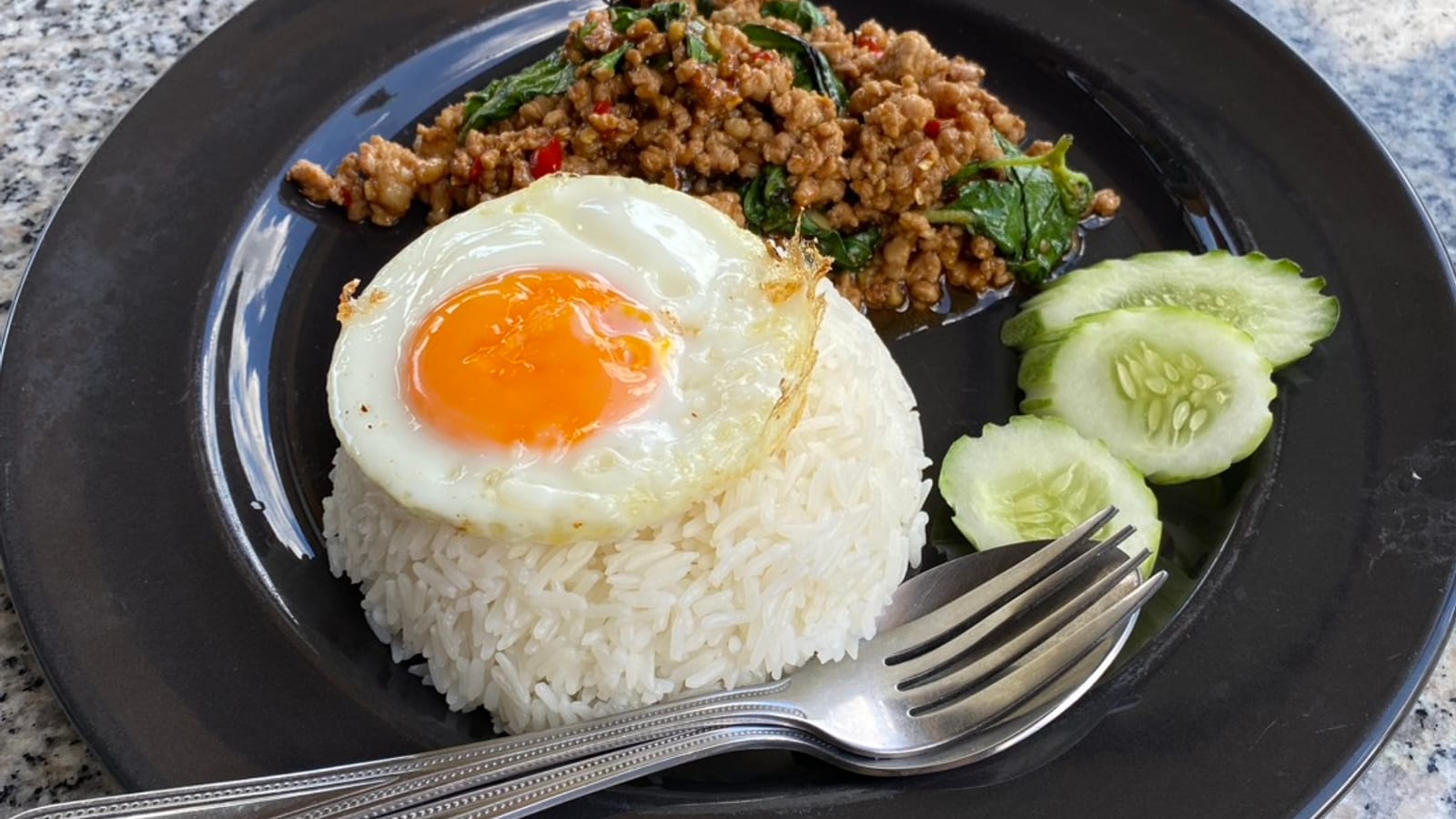 A single plate of pork gaprao — minced pork with basil and chilli — beside a mound of rice topped with a fried egg, three slices of cucumber, and one fork and spoon, on a granite table in daylight