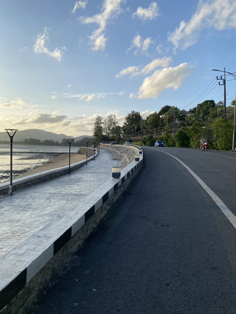 An empty concrete seafront promenade running beside a road in late afternoon, low tide exposing dark mudflats, hills across the water, and nobody on the walkway