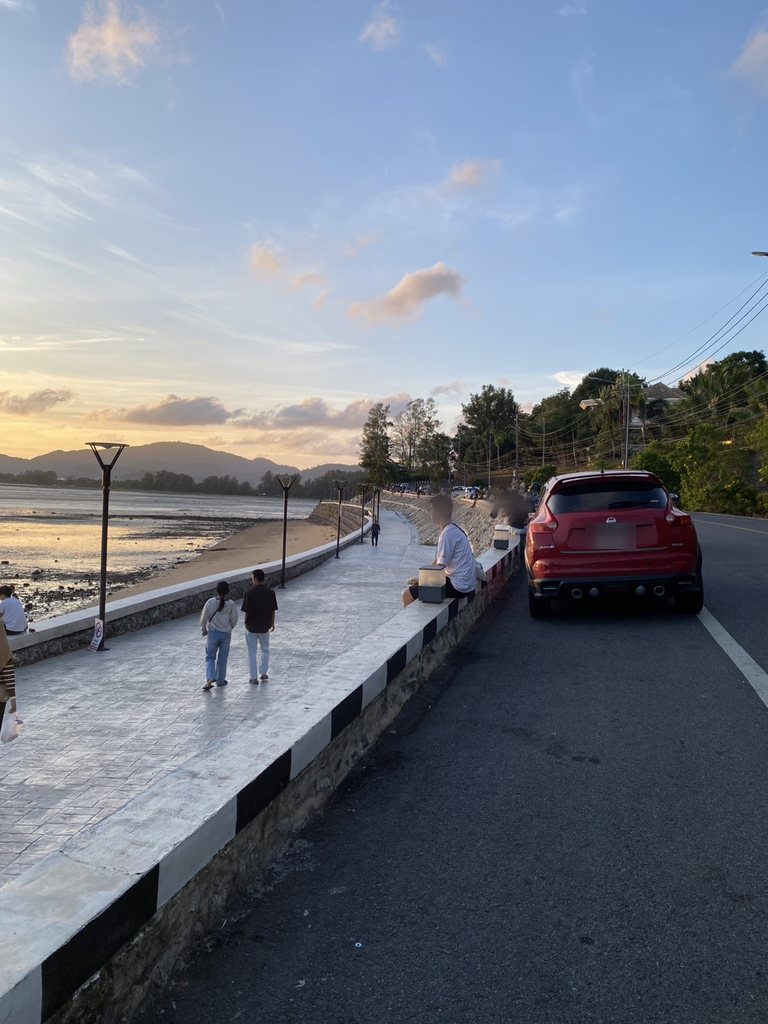 The same seafront promenade about an hour later in golden light, with people sitting along the sea wall, a couple walking, a car parked at the kerb and more cars lining the road into the distance