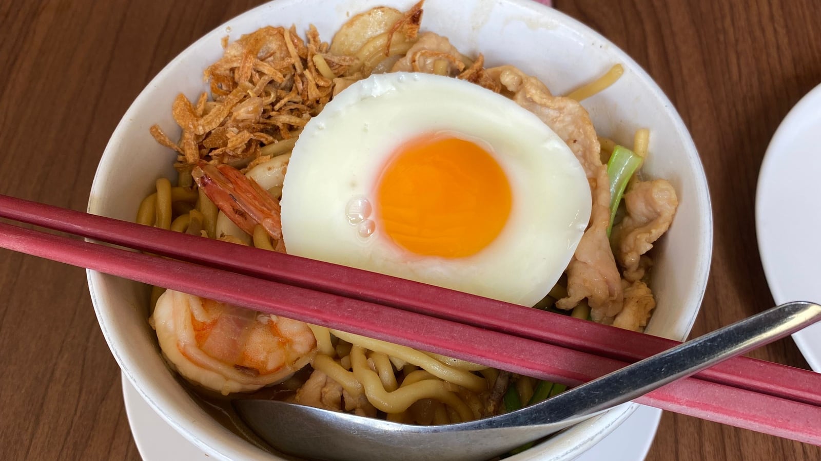 A single bowl of Phuket Hokkien mee — thick stir-fried yellow noodles with prawn, pork and a fried egg on top — with one pair of chopsticks and a spoon resting across it