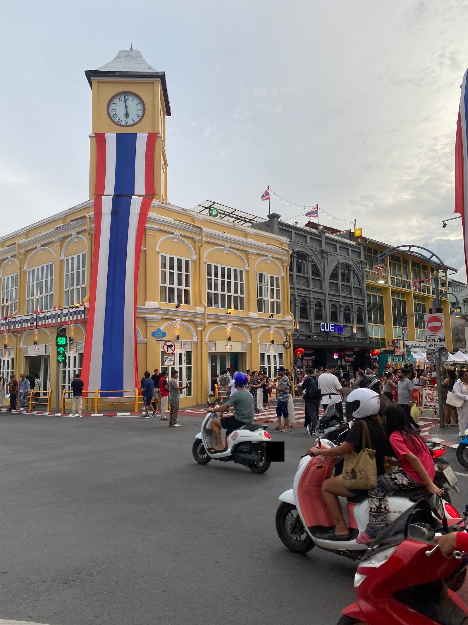 The clock tower at the edge of Phuket Old Town, draped in a Thai flag, with scooters crossing the junction in front of it and a crowd of people on the pavement outside the shophouses