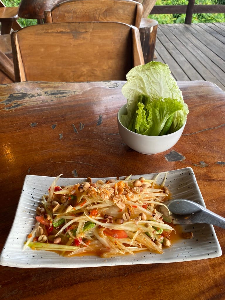 A plate of som tam with peanuts and a bowl of fresh cabbage and lettuce, on a worn wooden table on a deck