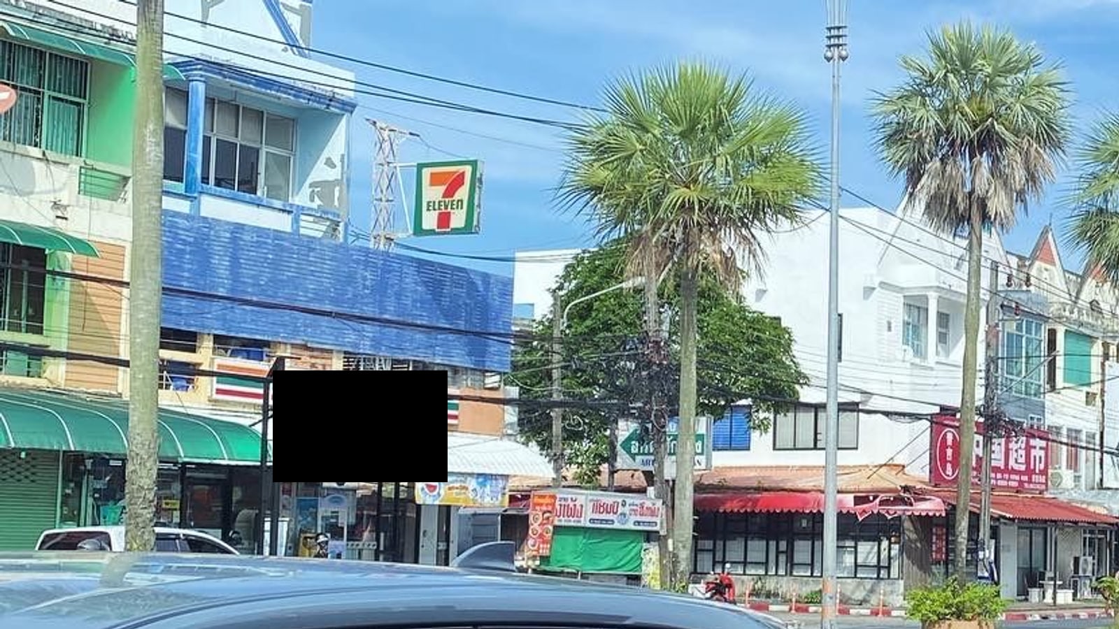 An ordinary street of shophouses in Phuket Town — green awnings, a 7-Eleven, sugar palms and overhead power lines under a bright sky
