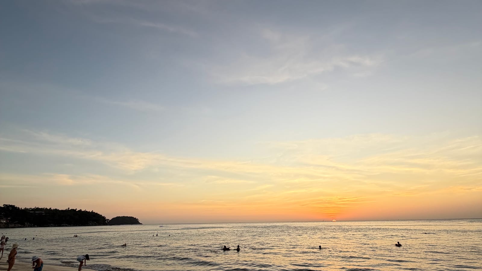 Karon beach at sunset, looking south — the sun just above the horizon over the Andaman, the wooded headland that separates Karon from Kata at the left with buildings among the trees, the islet of Koh Pu beyond its tip, people swimming in calm water, and footprints across wet sand in the foreground